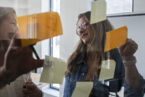 Deux femmes derrière une vitre collant des post-it pour un projet de stratégie salarié ambassadeur pour un client We Feel Good