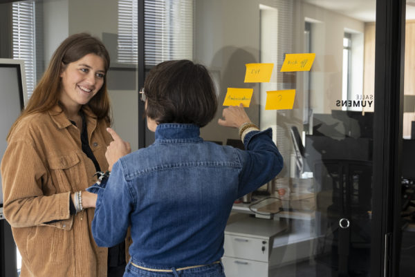 Deux femmes discutent et posent des post-it sur un mur vitré dans l'agence We Feel Good